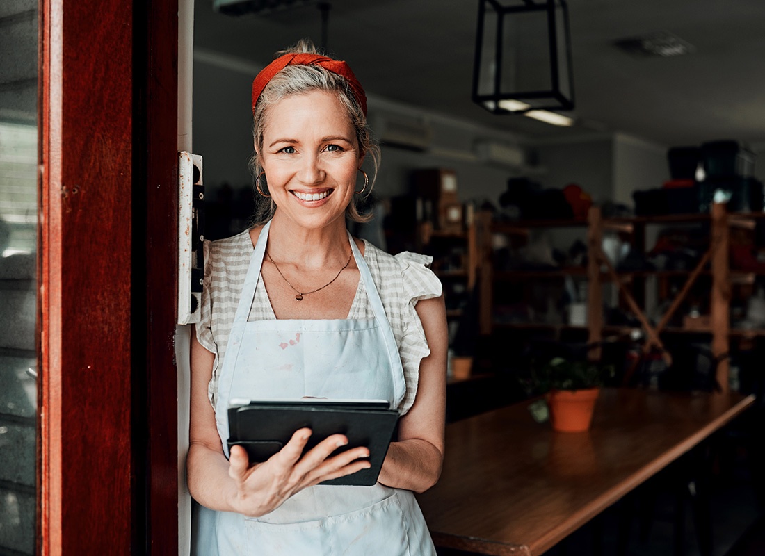 Business Insurance - Portrait of a Smiling Mature Business Woman Wearing an Apron Standing in the Front Entrance of her Main Street Shop While Holding a Tablet in her Hands
